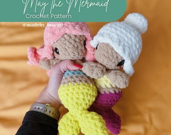 May include: Crocheted mermaid dolls in pink, white, and yellow. The dolls have brown skin and are held in a person's hand. The text "May the Mermaid Crochet Pattern" is in the background.