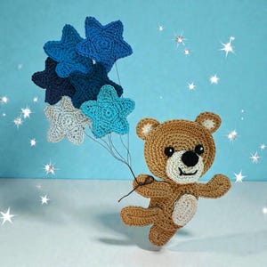 May include: A crocheted teddy bear with a smiling face, holding a bundle of star-shaped balloons in shades of blue. The bear is brown with a white muzzle and ears. The background is a light blue with sparkling star accents.