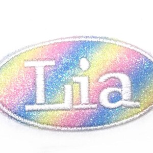 Puede incluir: Un parche ovalado blanco con un fondo de purpurina arcoíris y el nombre "Lia" escrito en blanco.