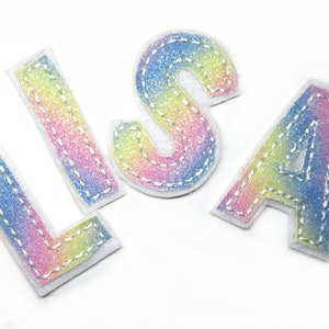 Könnte beinhalten: Vier Stoffbuchstaben mit dem Wort "LISA" in einem Regenbogen-Glitzer-Design. Jeder Buchstabe ist mit weißen Nähten umrandet und hat eine weiße Filzrückseite. Die Buchstaben sind in einer serifenlosen Schriftart und vor einem weißen Hintergrund angeordnet.