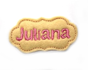 May include: A yellow felt name tag with a pink embroidered name "Juliana" in a decorative font.