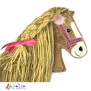 May include: A brown plush horse head with a braided yellow mane and tail. The horse has a pink glitter bridle and a pink flower on its head.
