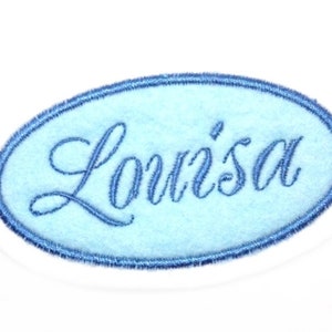 Könnte beinhalten: Ein hellblauer, ovaler Stoff-Patch mit weißem Rand und dem Namen "Loui'sa", der in blauer Farbe gestickt ist.