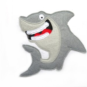 May include: A gray felt shark with a big, toothy grin and a red mouth. The shark is stitched with white thread and has a white eye.