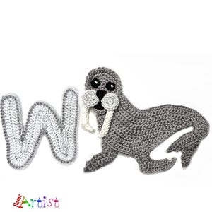 May include: A crocheted grey walrus with white tusks and a black face, beside a crocheted letter "W" with a grey outline. The walrus is lying down. The word "Home Artist" is in the bottom left corner.