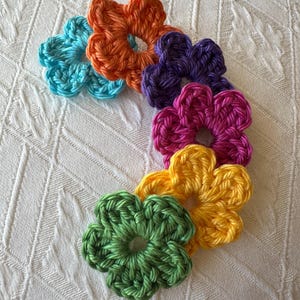 Könnte beinhalten: Eine Sammlung von sieben handgefertigten Häkelblumen in verschiedenen Farben, darunter Türkis, Orange, Lila, Pink, Gelb und Grün. Die Blumen sind auf einer weißen, strukturierten Oberfläche angeordnet. Jede Blume hat eine kreisförmige Mitte und blütenblattartige Ränder.