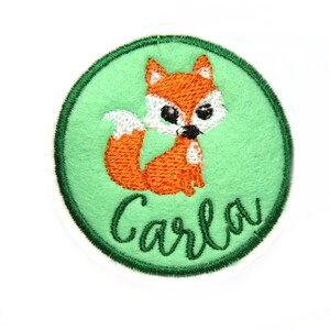 May include: A green circle with a white outline and a cute orange fox with a white tail and black eyes. The name "Carla" is embroidered in green below the fox.