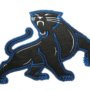 May include: A black panther patch with blue and white accents. The panther is in a roaring pose, with its mouth open and teeth visible. The patch is likely made of felt or a similar fabric, and is designed to be sewn or ironed onto clothing or accessories.