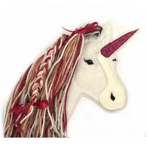 Könnte beinhalten: Weißer Filz-Einhornkopf mit einem rosafarbenen Glitzerhorn und einer geflochtenen Mähne aus rosa, braunem und weißem Garn. Das Einhorn hat ein rotes Band in seiner Mähne.