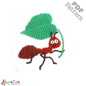 Puede incluir: Una hormiga roja de crochet con rayas marrones y una hoja verde. La hormiga tiene una cara sonriente con ojos blancos. La hoja está unida a la espalda de la hormiga con un hilo verde.