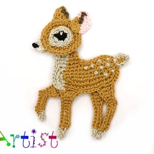 Puede incluir: Un cervatillo de crochet hecho con hilo marrón y beige, orejas rosas y ojos negros. El cervatillo está de pie sobre un fondo blanco.