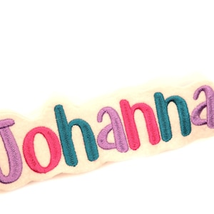 Könnte beinhalten: Ein weißer Filz-Patch mit dem Namen "Johanna", der in lila, rosa, türkis und grün gestickt ist.