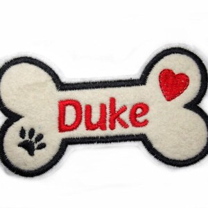 Könnte beinhalten: Weißer Filzhundeknochen mit schwarzem Umriss, rotem Herzen und schwarzem Pfotenabdruck. Der Name "Duke" ist in Rot gestickt.
