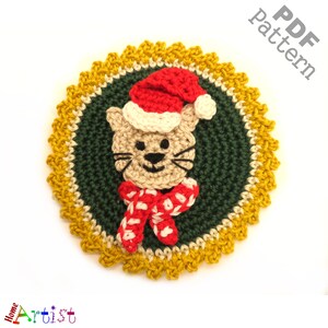 May include: Crocheted Christmas ornament featuring a cat wearing a red and white scarf and a Santa hat. The ornament is surrounded by a yellow and green crocheted border.