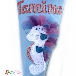 May include: A light blue cone-shaped item with the name "Tamina" in glittery pink letters. It features a white seahorse with blue accents and a pink and purple tulle mane. The bottom left corner has the word "Home Artist".