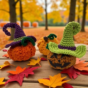 Puede incluir: Tres calabazas de ganchillo con sombreros de bruja y caras talladas sobre una superficie de madera con hojas de otoño coloridas. Las calabazas son naranjas, amarillas y marrones, con sombreros verdes y morados. El fondo está borroso.