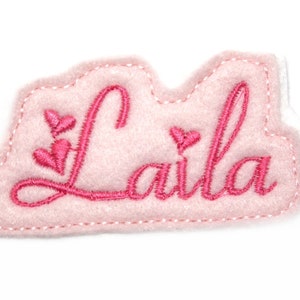 以下が含まれることがあります： ピンクのフェルト生地に「Laila」という名前がピンクの糸で刺繍されたアップリケ。アップリケは雲のような形をしていて、左側に小さなハートが3つ刺繍されています。