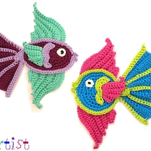 Könnte beinhalten: Zwei gehäkelte Fisch-Ornamente, eines mit einem Farbschema aus Lila, Grün und Blau und das andere mit einem Farbschema aus Rosa, Blau und Grün.