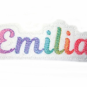 Può includere: Un toppa di feltro bianco con il nome "Emilia" ricamato in colori arcobaleno.