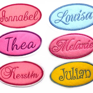 Könnte beinhalten: Sechs ovale Stoff-Patches mit gestickten Namen: Annabel, Louisa, Thea, Melanie, Kerstin und Julian. Die Patches sind in verschiedenen Farben, darunter rosa, blau, gelb und lila.