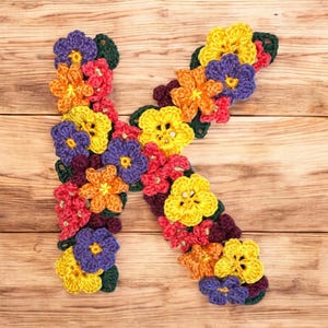 Può includere: La lettera K è realizzata con fiori all'uncinetto in tonalità di giallo, arancione, rosso, viola e verde. I fiori sono disposti in uno schema che ricorda la lettera K. Lo sfondo è una superficie di legno.