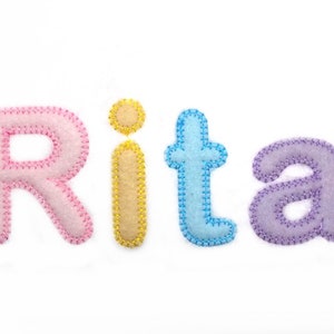 Könnte beinhalten: Der Name "Rita" in Filzbuchstaben geschrieben. Die Buchstaben sind rosa, gelb, blau und lila.