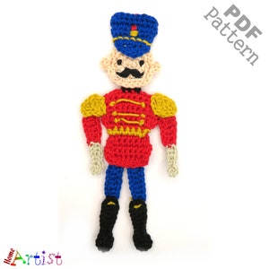 May include: Crochet pattern for a toy soldier. The soldier is wearing a red and yellow uniform, a blue hat, and black boots. The pattern is available as a PDF download.