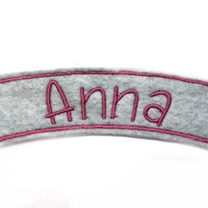 May include: A gray felt name patch with the name "Anna" embroidered in pink. The name is centered within a pink stitched border. The patch has a curved shape and is set against a white background.