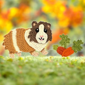 Op de afbeelding: Een gehaakt caviafiguurtje van bruin, beige en crèmekleurig garen, met zwarte veiligheidsoogjes en een roze geborduurde neus. Naast de cavia ligt een kleine gehaakte oranje wortel met groen loof gemaakt van garen.