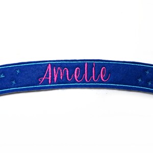 Könnte beinhalten: Ein blaues Filzbanner mit dem Namen "Amelie" in rosa Schrift gestickt. Das Banner hat einen hellblauen Rand und kleine sternförmige Verzierungen. Ein dekorativer Artikel.