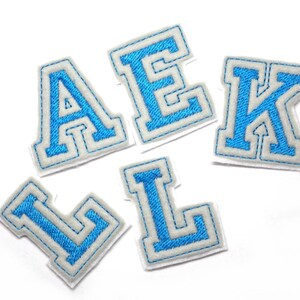 May include: Five embroidered felt letter patches in blue and white. The letters are A, E, K, L, and L. Each letter has a blue center with a white border and a gray outline. The patches are on a white background.