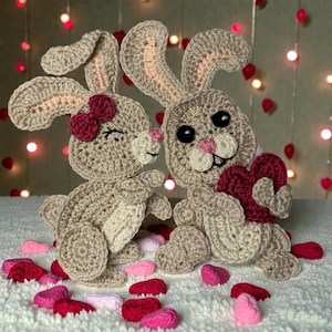 May include: Two crocheted bunnies in a loving embrace. One bunny has a red bow and closed eyes, the other holds a red heart. The bunnies are beige with pink accents, surrounded by red and pink heart-shaped decorations.