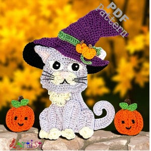 May include: Crocheted Halloween cat wearing a purple witch hat with orange pumpkin accents. Two crocheted orange pumpkins with green stems flank the cat. The cat is light gray with black whiskers and eyes. The background is a blurred yellow and orange.