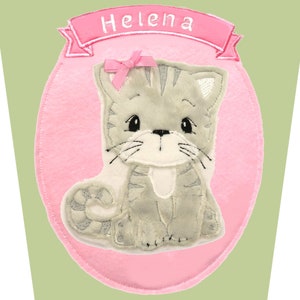 May include: A gray and white cat applique with a pink bow and the name "Helena" on a pink banner. The cat is set against a pink oval on a light green background. The cat has a cute, cartoonish design.