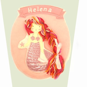 May include: Pink felt mermaid applique with a pink, orange, red, and white yarn braid. The mermaid has a pink glitter tail and a pink seashell bra. The applique has the name "Helena" written on a pink banner.