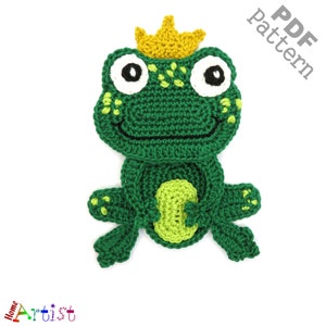 May include: Green crocheted frog wearing a yellow crown. The frog has black eyes and a smiling mouth. The frog is sitting on a white background.