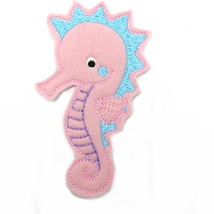 Peut inclure: Hippocampe en feutre rose avec des broderies bleues et violettes. L'hippocampe a une grande nageoire sur la tête et une queue enroulée.