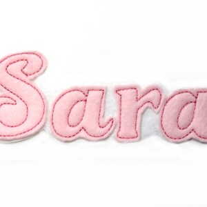 Könnte beinhalten: Rosa Filzbuchstaben, die den Namen "Sara" in einer dekorativen Schreibschrift buchstabieren. Die Buchstaben sind mit rotem Faden gestickt.