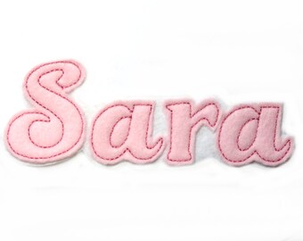 May include: Pink felt letters spelling out the name "Sara" in a decorative script font. The letters are stitched with red thread.