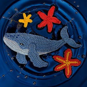May include: A crocheted blue whale swims among red, yellow, and orange starfish in a blue water setting. The whale has a light blue belly and a dark blue back. The starfish are in various shades of red, yellow, and orange.
