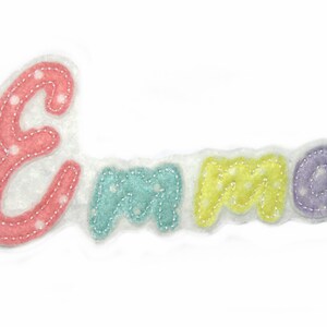 Könnte beinhalten: Eine Filzapplikation des Namens "Emma" in Pastellfarben mit weißen Punkten. Die Buchstaben sind mit weißen Nähten umrandet.