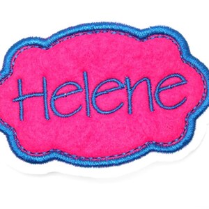 Könnte beinhalten: Rosa Filznamensschild mit blau besticktem Rand und dem Namen "Helene" in blauer Farbe.