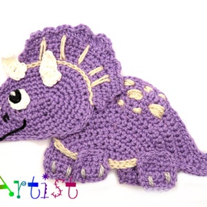 May include: A handmade crochet triceratops dinosaur in shades of purple, cream, and black. The dinosaur features three horns, a frill, and a friendly expression. The text "Home Artist" is visible in the lower left corner.