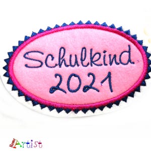 May include: A pink oval patch with the German word "Schulkind" and the year "2021" embroidered in dark blue. The patch has a dark blue and white scalloped border and a pink inner border.
