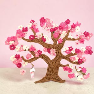 May include: A crocheted tree sculpture featuring a brown yarn trunk and branches adorned with numerous small, five-petaled flowers in various shades of pink, magenta, and white, creating a dense, blooming canopy effect across the structure.
