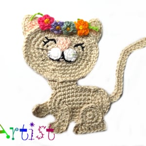 May include: A crocheted cat with a flower crown. The cat is beige and has a white face with black eyes and whiskers. The flower crown is made of pink, purple, orange, and green yarn.