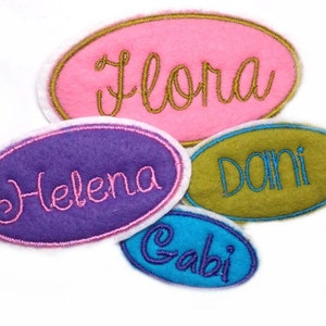 Puede incluir: Cuatro etiquetas de nombre de fieltro de forma ovalada con nombres bordados. Los nombres son "Flora", "Helena", "Dani" y "Gabi". Las etiquetas son de color rosa, morado, verde y azul.