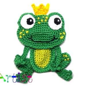May include: A green crocheted frog wearing a yellow crown. The frog has white eyes with black pupils and a black smile. The frog is sitting with its legs outstretched.
