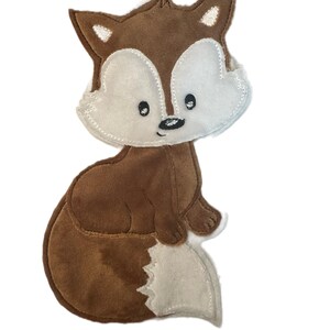 Könnte beinhalten: Eine braun-cremefarbene Applikation aus Stoff in Fuchsform mit gestickten Details. Der Fuchs hat einen dunkelbraunen Körper, ein cremefarbenes Gesicht und eine cremefarbene Schwanzspitze, schwarze Augen mit weißen Akzenten und eine kleine Nase.