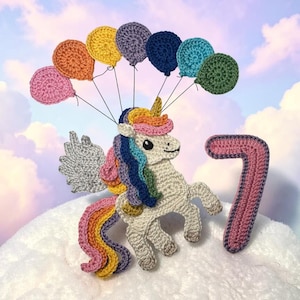 May include: A crocheted unicorn with a rainbow mane, tail, and wings, holding a string of colourful balloons. A crocheted number 7 is next to the unicorn. The background is a cloudy sky.